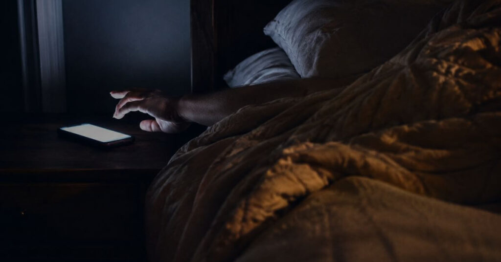 A dim bedroom in pre-dawn stillness, a glowing iPhone on a nightstand illuminating an outstretched arm; featured image for the poem Ten Sacred Minutes on Sayspire.